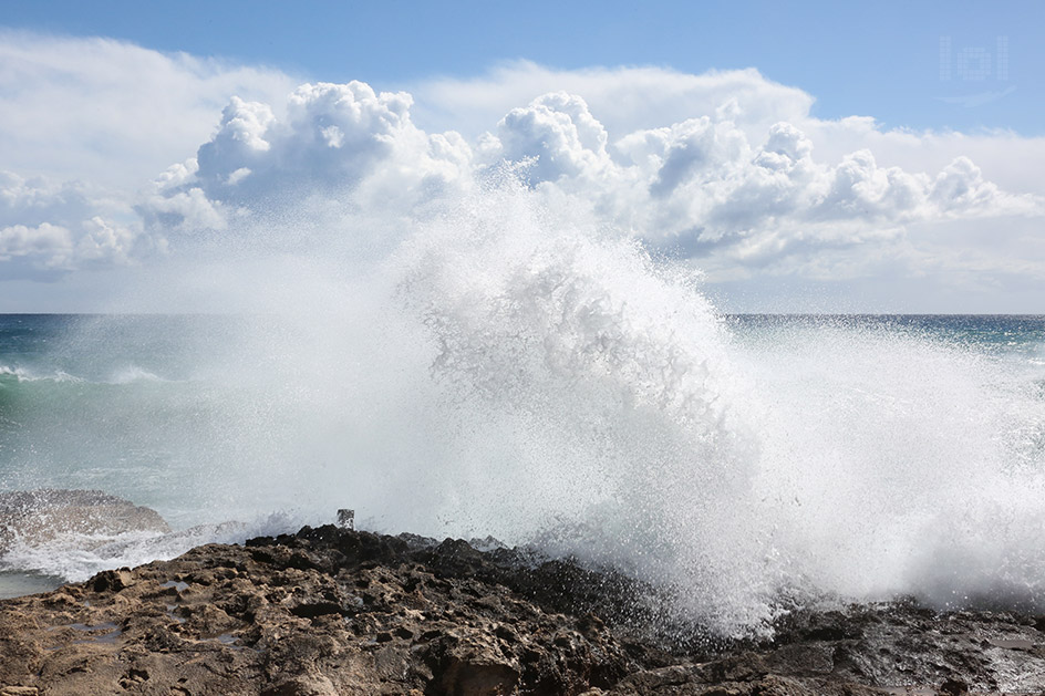 Große Welle bricht mit voller Wucht am Felsen, dahinter eine dramatische Wolke